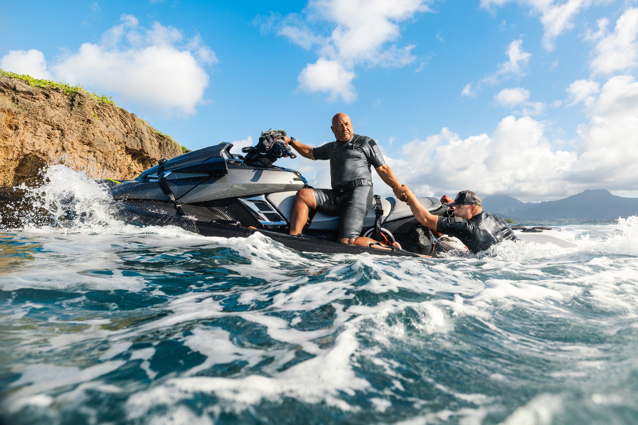 Two men wearing Waihana Tropicam Black Short Sleeve Spring Suits work together beside a jet ski in churning ocean water near a rocky Hawaiian coastline.